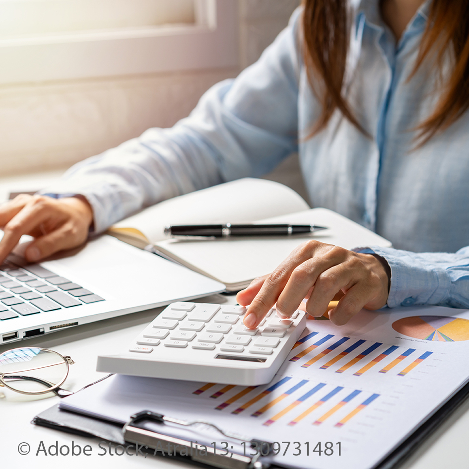 Eine Frau mit vor einem Notbook. Sie tippt auf einem Taschenrechner. Auf dem Tisch liegen statistische Grafiken.