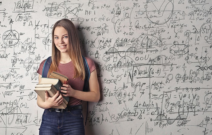 Eine junge Frau steht vor einer Wand mit Formeln und Grafiken. Sie hält Bücher in der rechten Hand und lächelt in die Kamera.