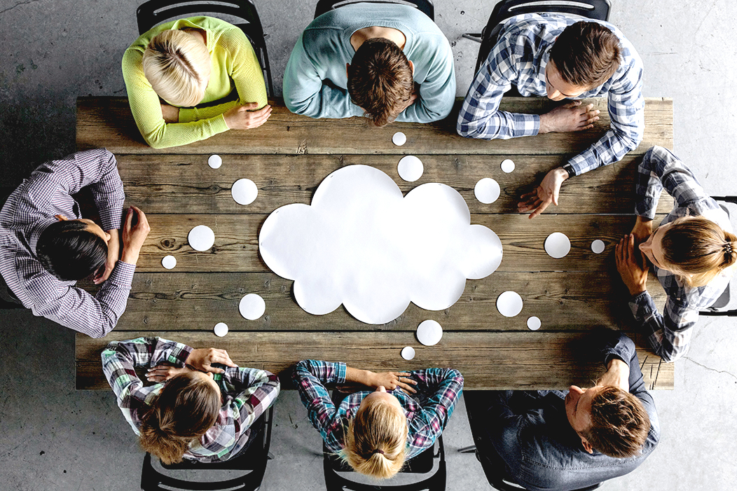 Bild aus der Vogelperspektive. Ein Team acht junger Menschen sitzt am Tisch. In der Mitte liegt eine weiße leere Papierwolke.