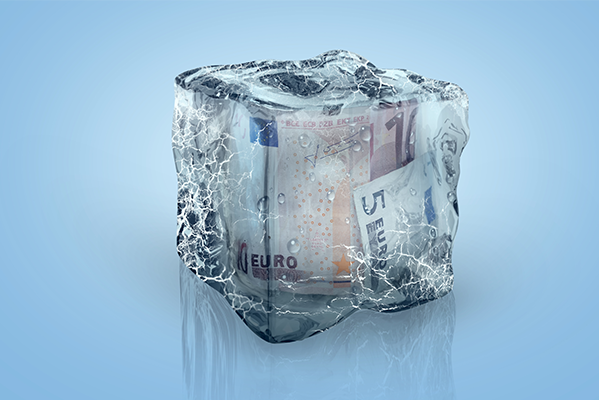 Ein Eiswürfel, in dem mehrere Euro-Banknoten eingeschlossen sind. vor einem blauen Hintergrund.