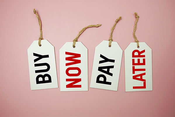 Vier Etiketten mit der Aufschrift "Buy now pay later" vor einem pinken Hintegrund. Jedes Wort ist einzeln auf einem Etikett.