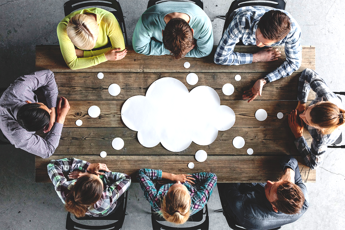 Bild aus der Vogelperspektive. Ein Team aus acht jungen Menschen sitzt am Tisch. In der Mitte liegt eine weiße leere Papierwolke.