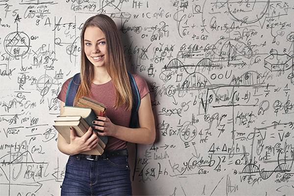 Eine junge Frau steht vor einer Wand mit Formeln und Grafiken. Sie hält Bücher in der rechten Hand und lächelt in die Kamera.