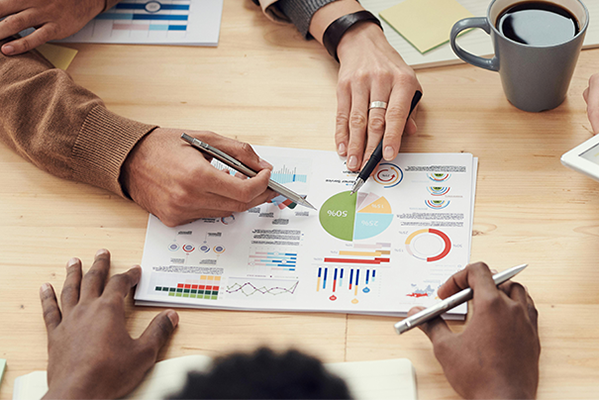 Ein Team von drei Personen an einem Tisch. Es sind nur ihre Hände zu sehen. Sie diskutieren über Grafiken und Zahlen.